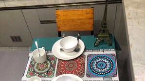 Uma mesa de jantar com toalha de mesa verde-água, posta com pratos, tigelas e talheres brancos. Um modelo da Torre Eiffel é visível ao fundo.