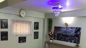 Uma cama bem arrumada com almofadas e uma toalha enrolada, um relógio de parede, quadros emoldurados, uma pintura grande de uma paisagem urbana e um candelabro.