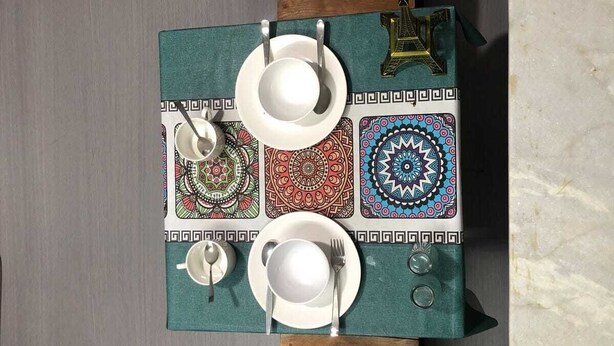 Uma área de jantar com uma mesa posta para quatro pessoas, com um jogo de toalha rendado e cadeiras de madeira.