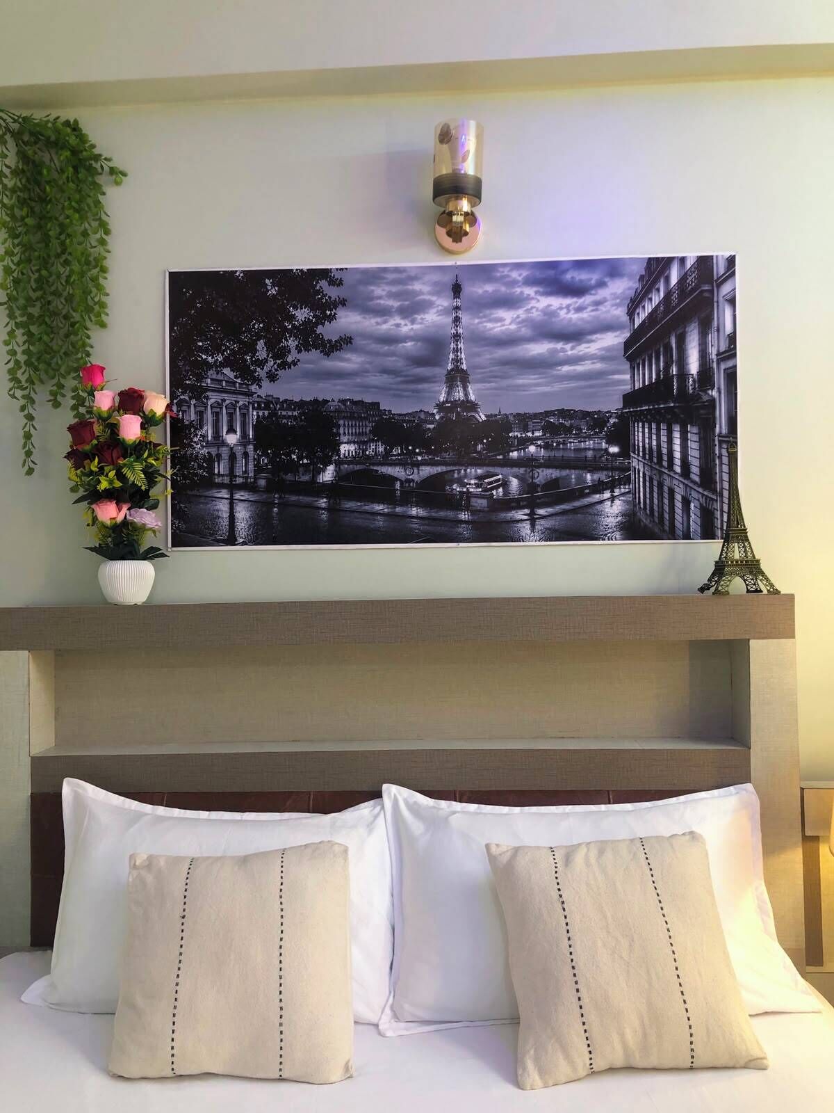 A bedroom with a bed, pillows, a framed picture of the Eiffel Tower, a small vase with flowers, and a wall-mounted light fixture.
