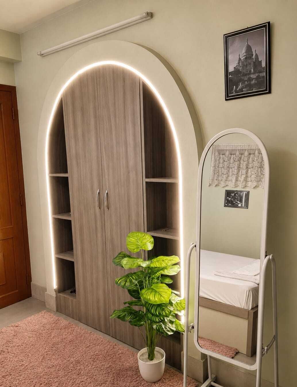 A room with a large arched wardrobe, a bedside table with a mirror, a potted plant, and a framed picture on the wall.