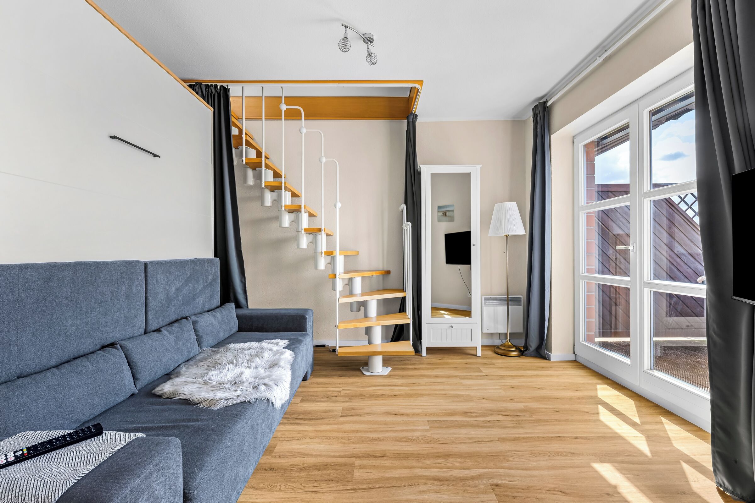 A modern living room with a grey sofa, wooden floor, a white staircase, and large windows.