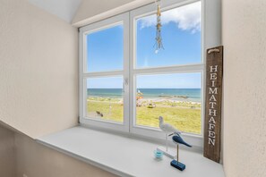 Una habitación con un amplio ventanal con vistas a una playa, un letrero de madera con el nombre 'HEIMATHEEN' y una figurita de pájaro en el alféizar de la ventana.