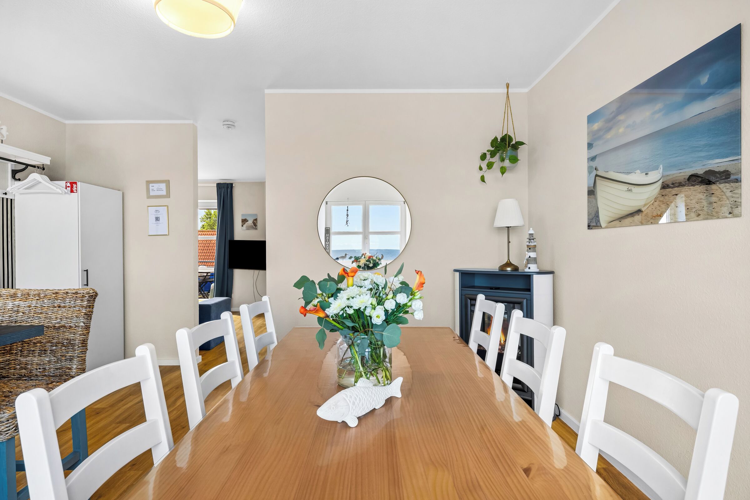 Et spiseområde med et træbord, hvide stole, en vase med blomster, et rundt spejl og et billede af en strand.