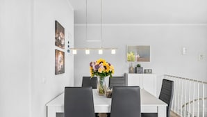 Uma sala de jantar moderna com uma mesa branca, cadeiras pretas, um vaso de flores e um corrimão branco ao fundo.