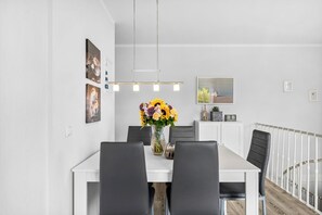 Un comedor moderno con una mesa blanca, sillas negras, un jarrón de flores y una barandilla blanca al fondo.