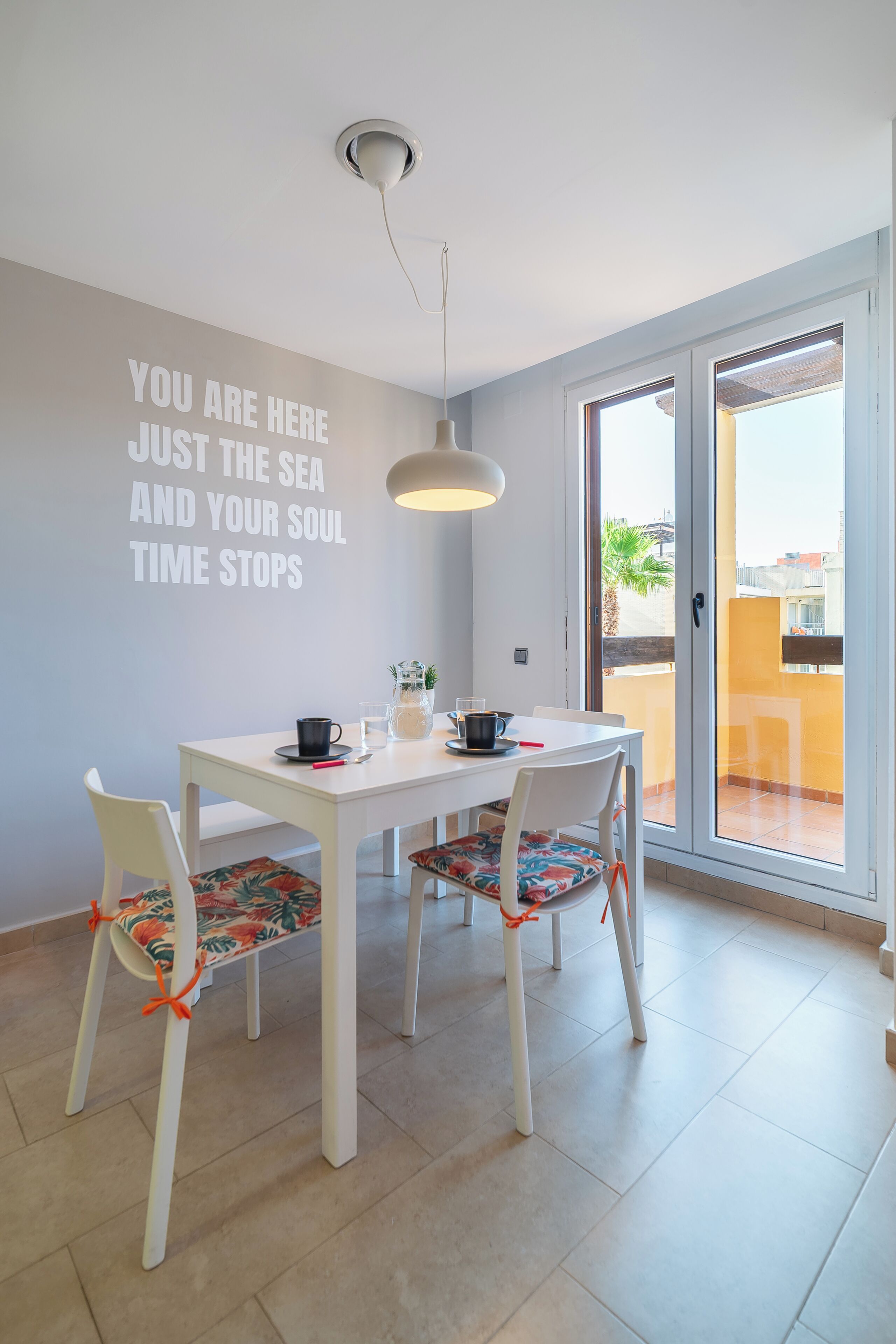 Un coin repas avec une table et des chaises blanches, un luminaire suspendu et un mur orné d’un texte motivant.