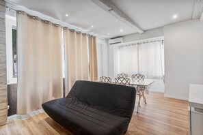 Una habitación moderna con un sofá negro, piso de madera, una mesa de comedor con sillas y un ventanal con cortinas.