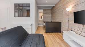 Un intérieur moderne avec un canapé, un téléviseur à écran plat fixé sur un mur en bois, et un coin cuisine visible à travers une porte en verre.