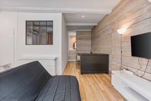 Un interior moderno con un sofá, un televisor de pantalla plana montado en una pared de madera, y una cocina visible a través de un vidrio.