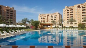 A hotel pool with lounge chairs and umbrellas, surrounded by palm trees and a clear sky.