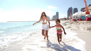 A beach scene with a city skyline in the background, including high-rise buildings and a clear sky.