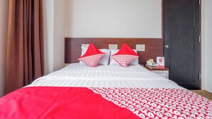 Ett hotellrum med en säng, två röda kuddar, ett vitt överkast, en röd pläd, ett träfärgat sänggavel, ett nattduksbord, en telefon och en dörr.