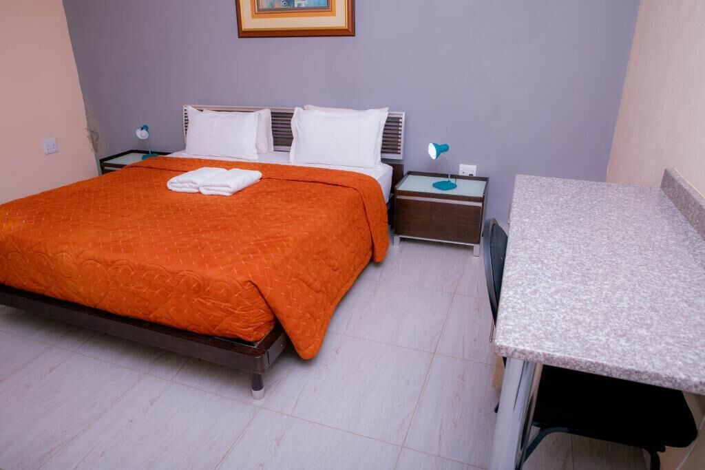 A hotel room with a bed, a desk, a chair, a nightstand, and a wall-mounted lamp.