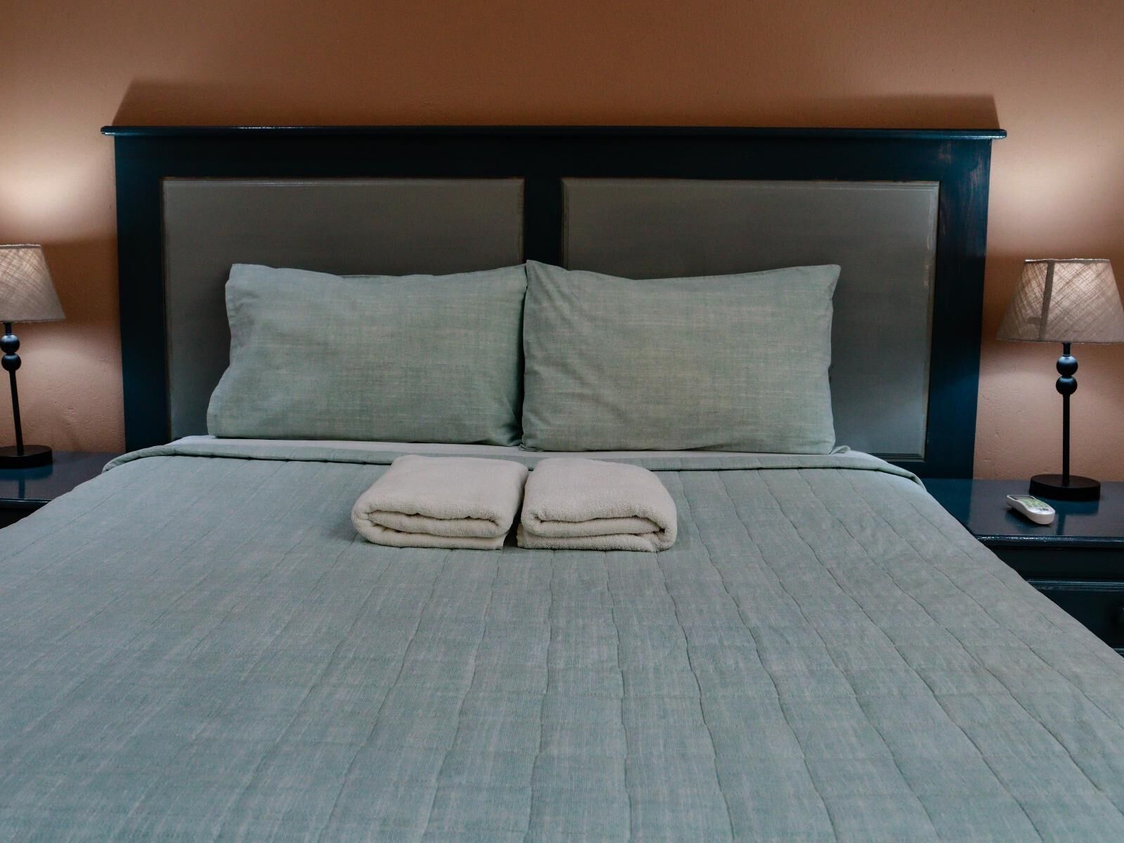 Uma cama bem arrumada com um edredom cinza e duas almofadas, um controle remoto na mesa de cabeceira e um abajur na mesinha lateral.