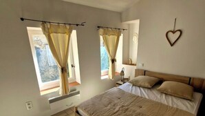 Een slaapkamer met een groot bed, twee ramen voorzien van gordijnen, een hartvormige wanddecoratie en een geweven mand.