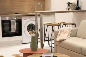 A modern kitchen with a built-in oven, washing machine, and a coffee machine on the countertop. There are stools and a sofa in the living area. - celine 16 (Paris)