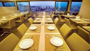 Un restaurante con varias mesas dispuestas para comer, organizadas en filas, y una vista de la ciudad visible a través de amplios ventanales.