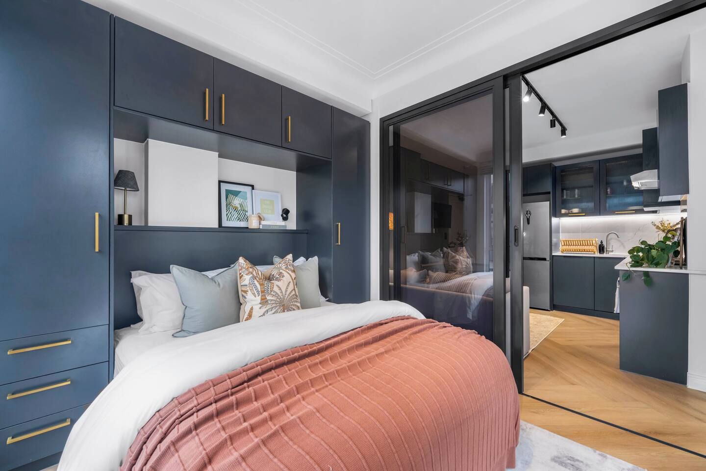Ein modernes Schlafzimmer mit einem großen Bett, einem Kleiderschrank mit goldenen Griffen und Blick in die Küche durch eine Glastür.