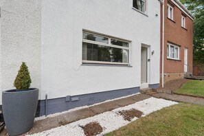 Ein zweistöckiges Haus mit weißer Fassade, ein kleiner Vorgarten mit weißen Kieselsteinen und ein hoher, schmaler Pflanzkasten.