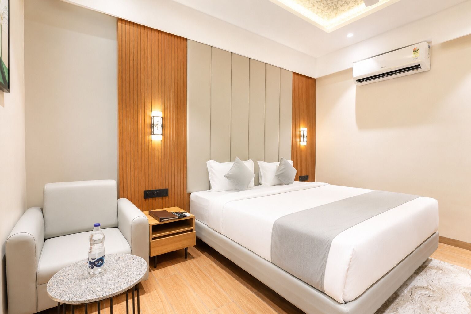 A hotel room with a bed, a seating area with a table, a water bottle, and an air conditioning unit.