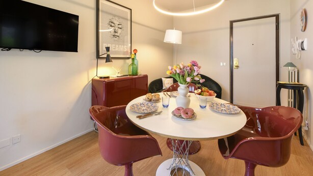 Et spiseområde med et rundt bord, røde stole, en vase med blomster og et fladskærms-tv på væggen.
