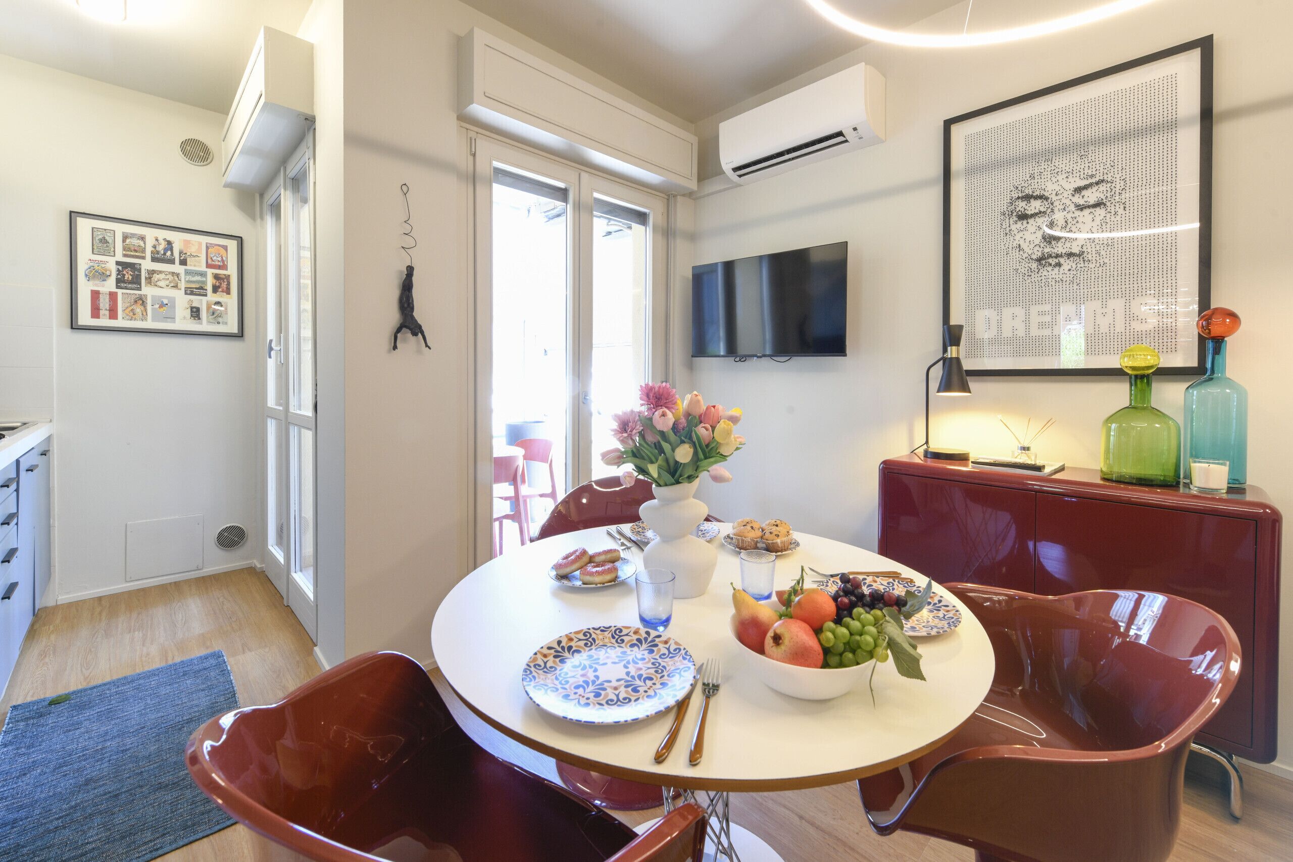 A dining area with a round table, chairs, a vase with flowers, and a fruit bowl.