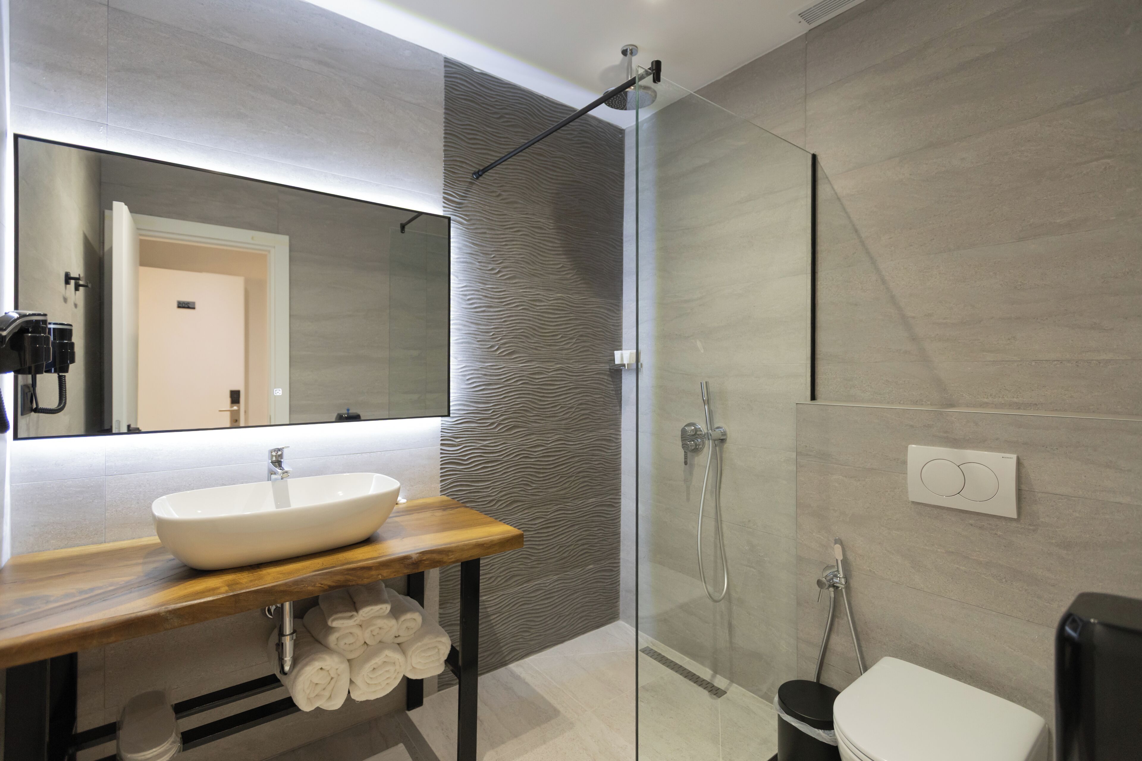Un baño moderno con ducha envuelta en vidrio, tocador de madera con lavamanos blanco y un espejo grande.