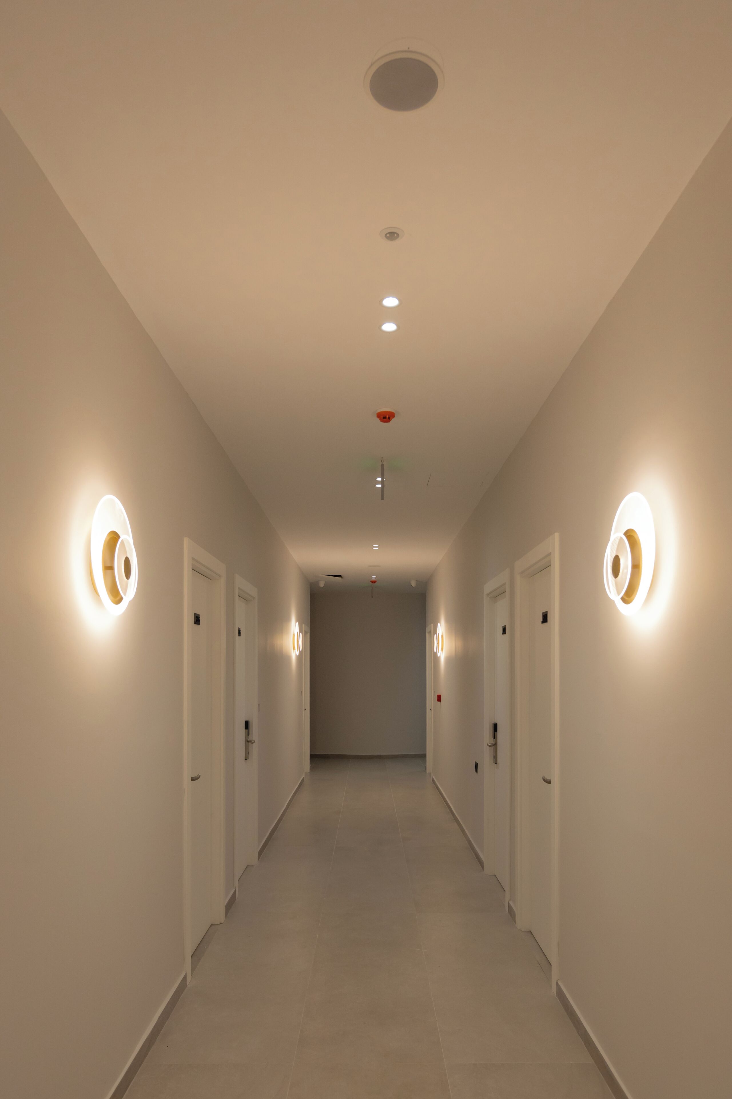 A hallway with white walls, doors on both sides, and circular light fixtures on the walls.