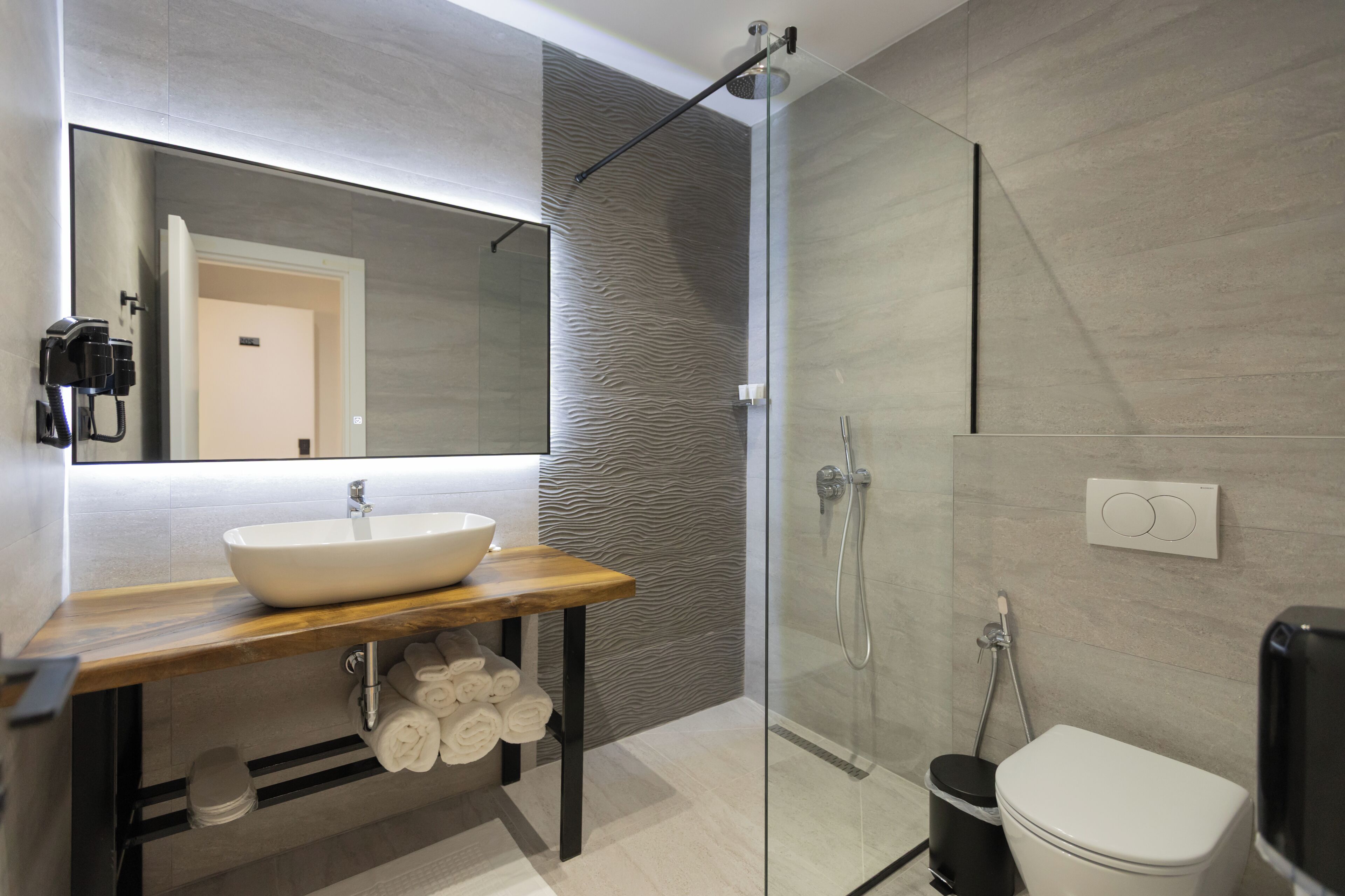 Un baño moderno con un espejo grande, un tocador de madera, un lavabo blanco, cabina de ducha de vidrio y un inodoro.