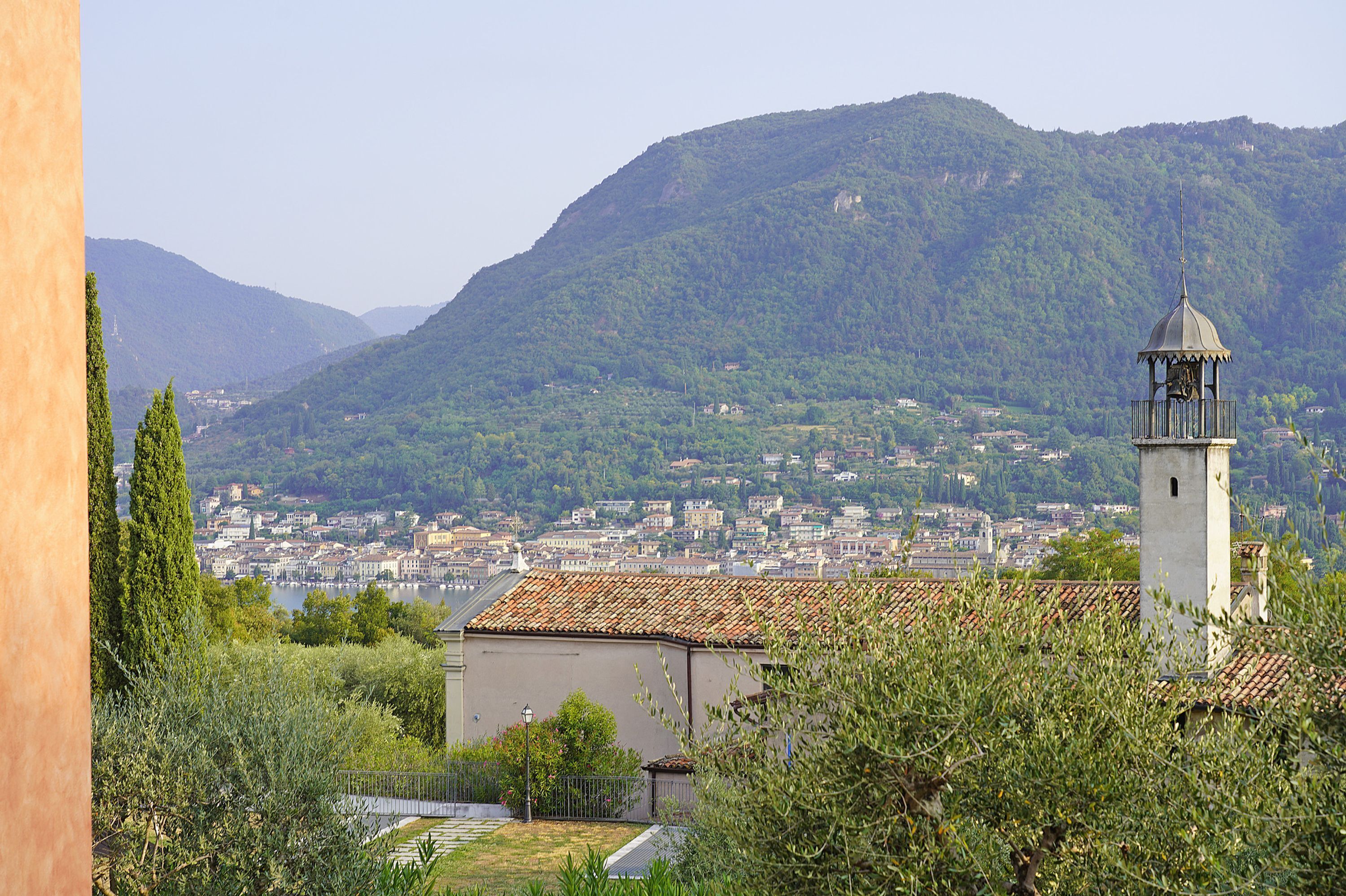 A view of a town with a prominent bell tower, surrounded by greenery and mountains.