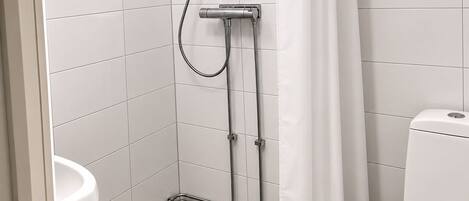 A bathroom with white tiles, a shower with a handheld showerhead, a toilet, and a white curtain.