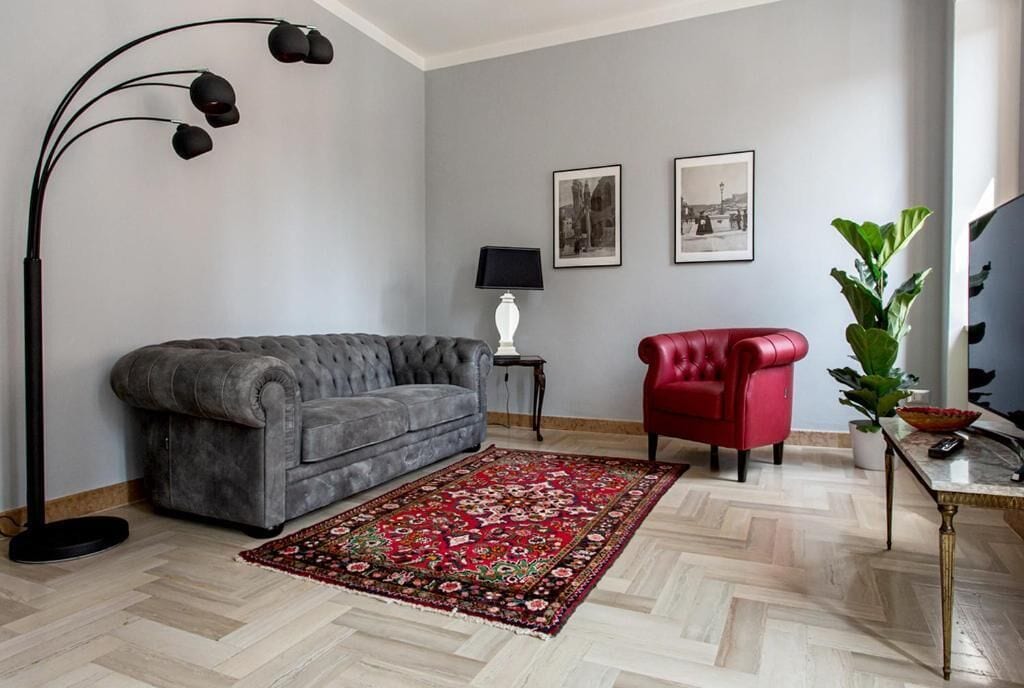 Sala de estar con un sofá gris, un sillón rojo, un tapete estampado, una lámpara de pie y dos cuadros en la pared.