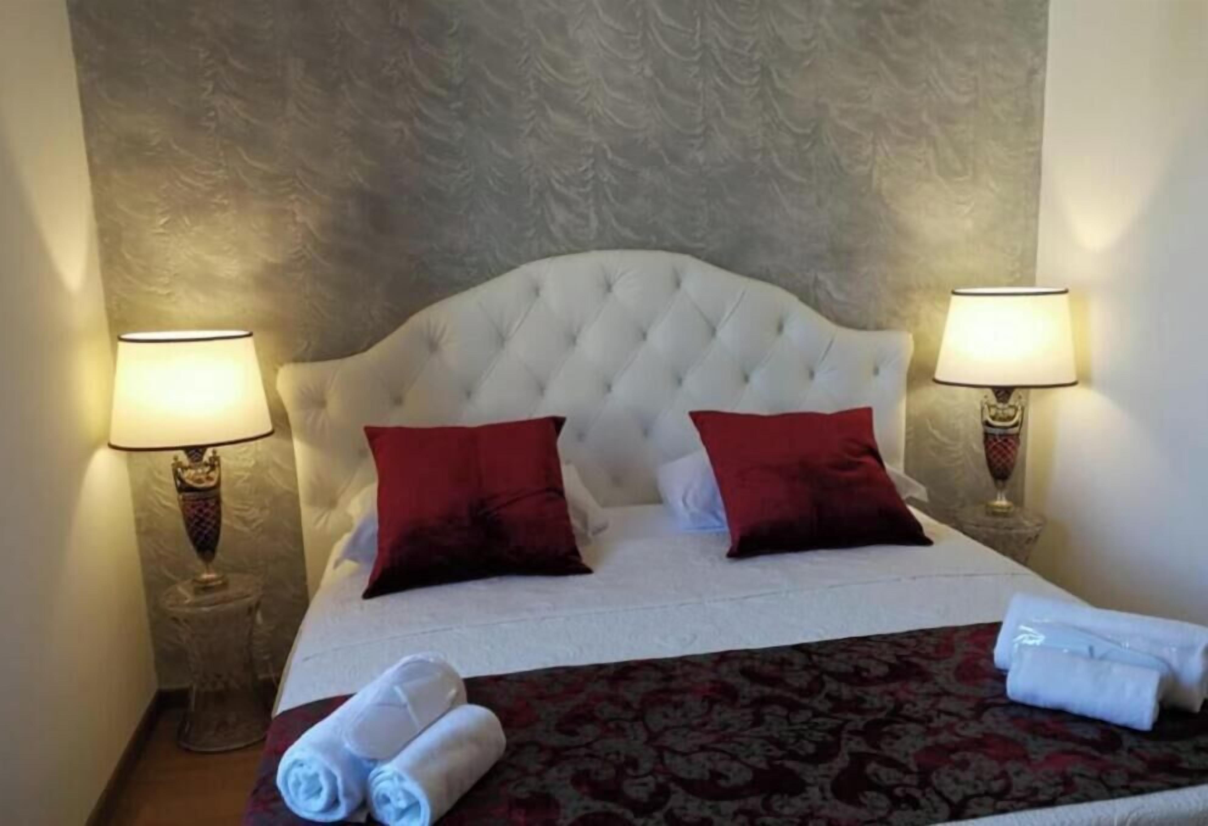 Una habitación de hotel con un cabecero capitonado, dos almohadas rojas, una colcha blanca, dos lámparas de noche y una toalla enrollada sobre la cama.
