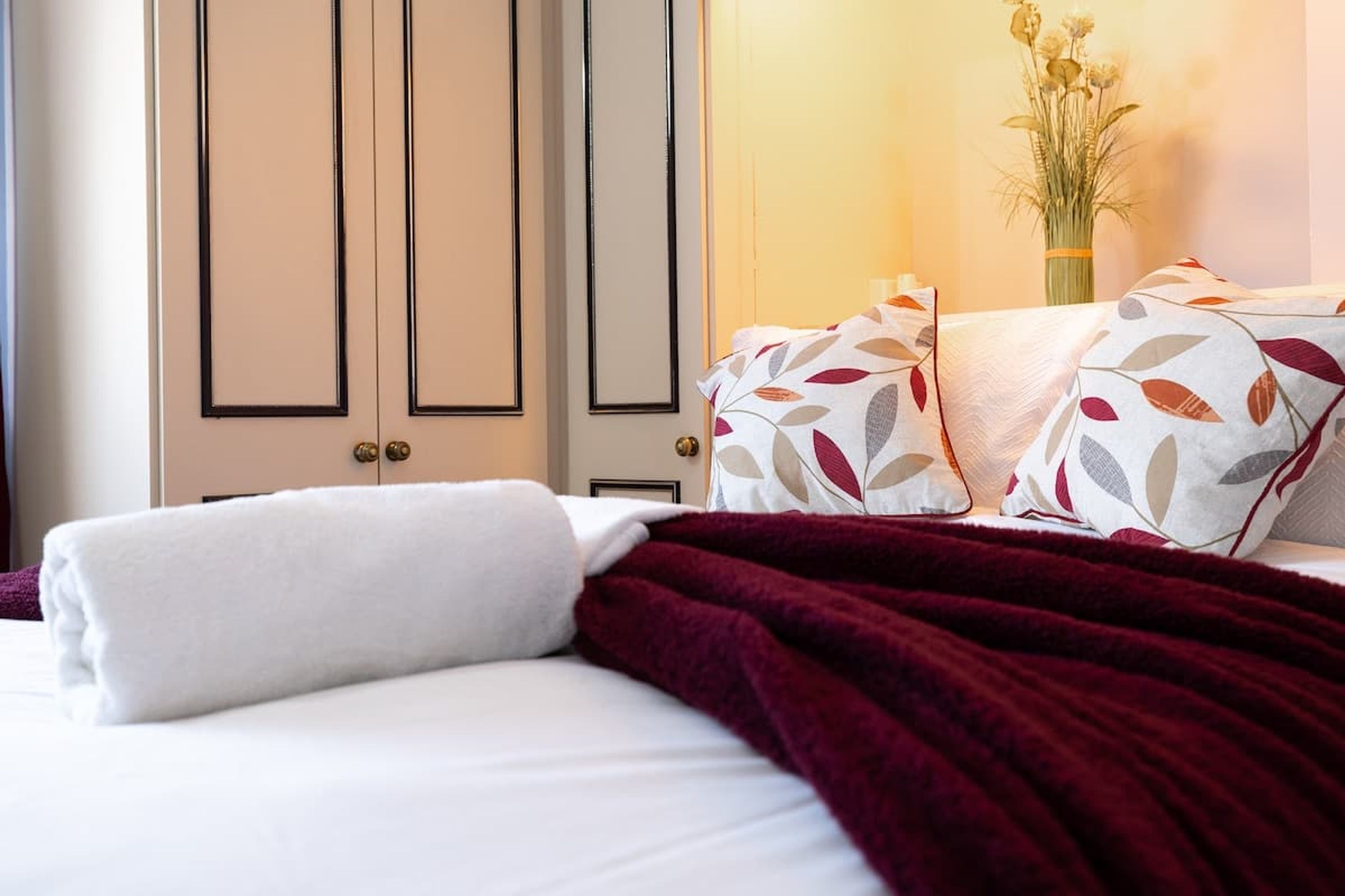 A hotel room with a bed, a red throw blanket, and a vase with flowers on a nightstand.