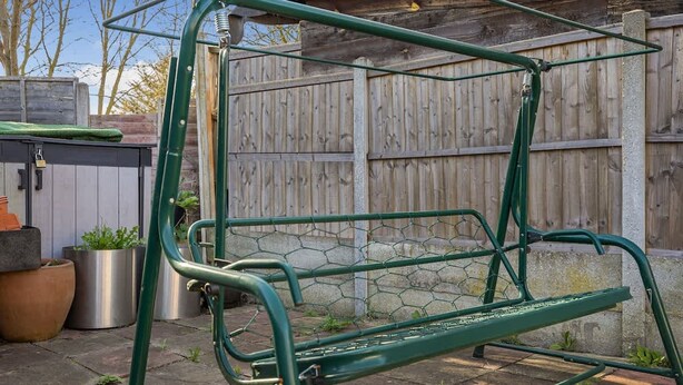 A green metal swing set in a backyard with a wooden fence, potted plants, and a stone wall.