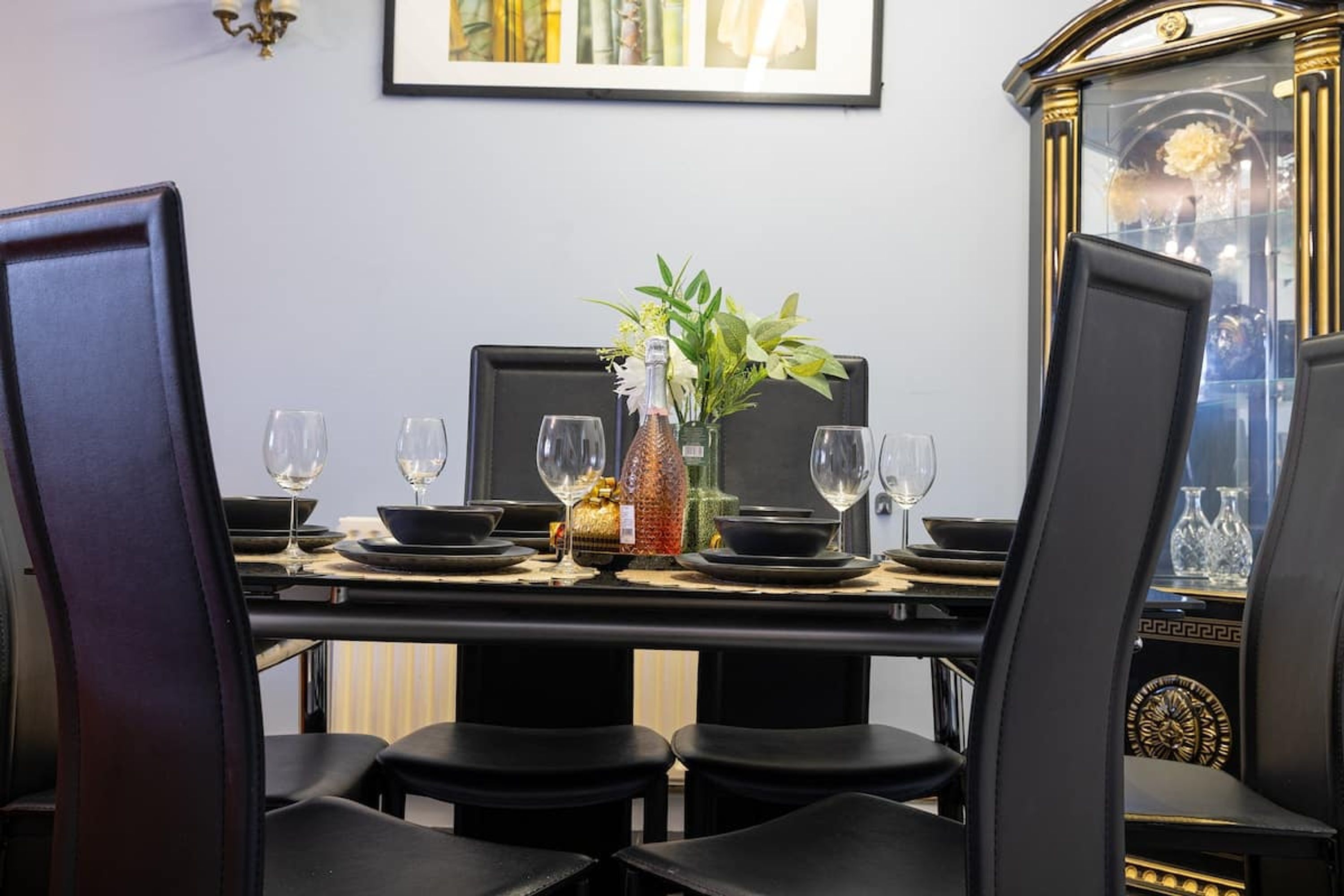 Una mesa de comedor para cuatro personas, con sillas negras, un florero con plantas verdes y copas de vino.