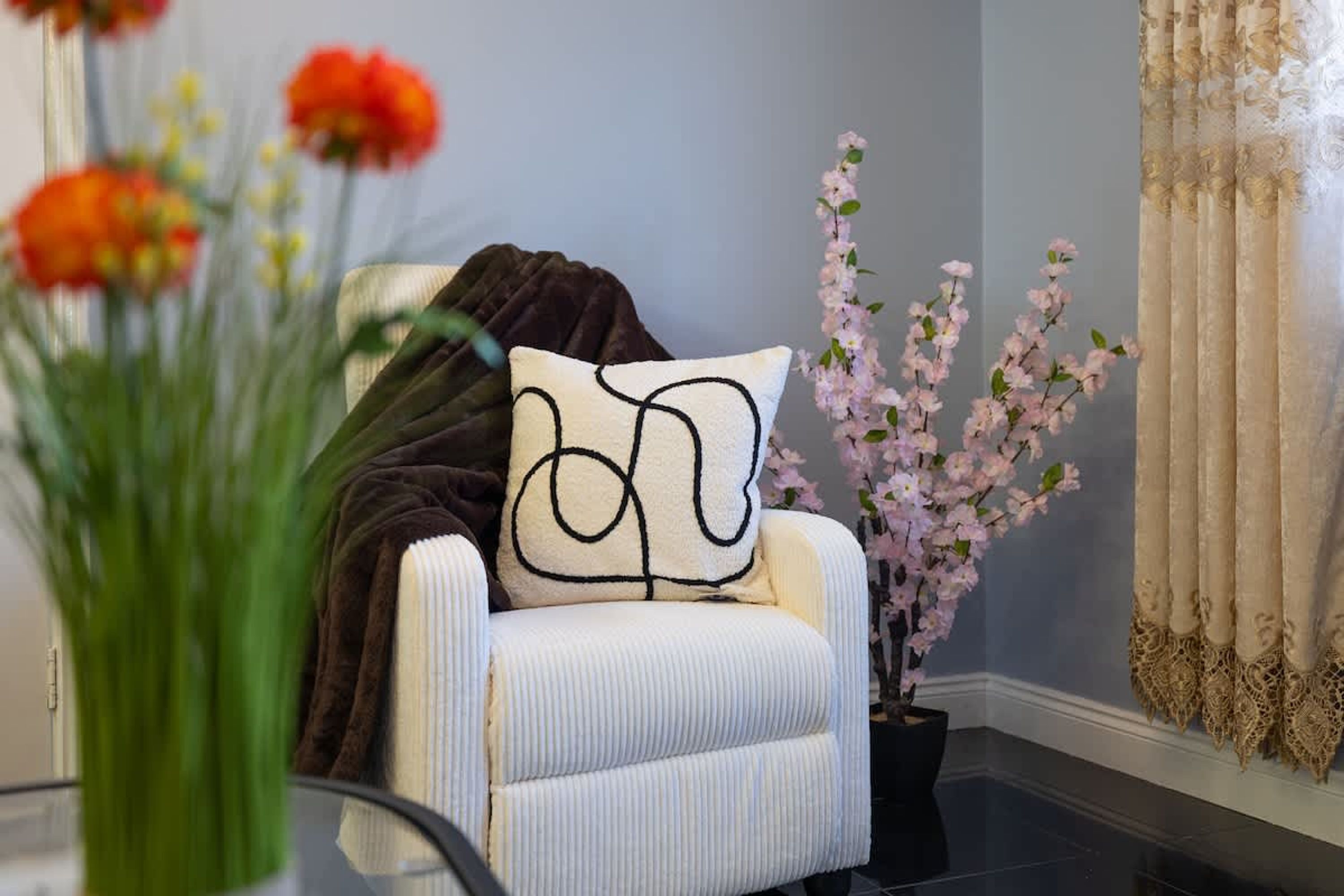 Una habitación con un sillón blanco, una manta negra, un florero con flores y una planta en maceta con florecillas rosas.