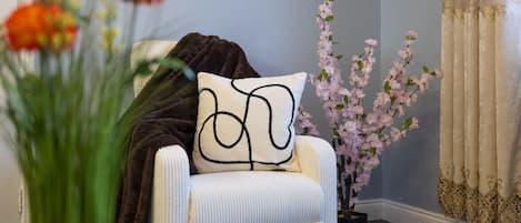 A room with a white armchair, a black throw blanket, a vase with flowers, and a potted plant with pink blossoms.