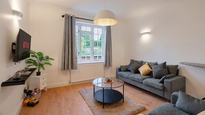 Ein modernes Wohnzimmer mit grauem Sofa, Couchtisch, Wandfernseher und einem Fenster mit Vorhängen.