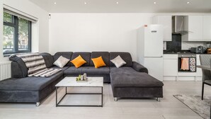 A modern living room with a sectional sofa, a coffee table, and a kitchen area with white cabinets and appliances. - Comfy 3BR House Near Tottenham Stadium Parking (London)