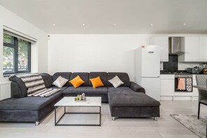 A modern living room with a sectional sofa, a coffee table, and a kitchen area with white cabinets and appliances. - Comfy 3BR House Near Tottenham Stadium Parking (London)