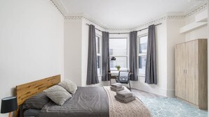 A bedroom with a bed, a nightstand, a lamp, a chair, a window with curtains, and a wardrobe. - Specious 2BR Family Home Parking Sleeps 6 (London)
