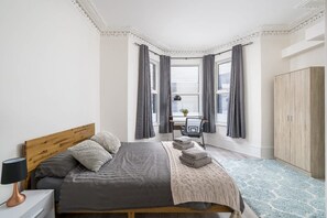 Ein Schlafzimmer mit einem Bett, einem Nachttisch, einer Lampe, einem Sessel, einem Fenster mit Vorhängen und einem Kleiderschrank.