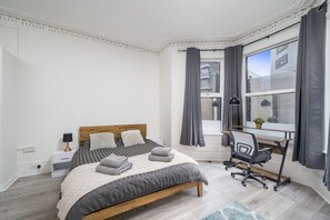 Ein Schlafzimmer mit Bett, Nachttisch, Lampe, Schreibtisch, Stuhl und Blick ins Freie.