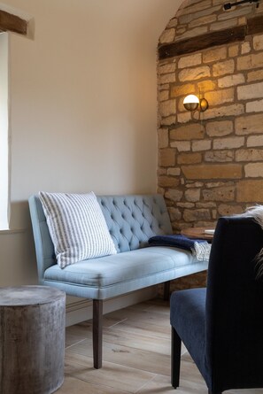 A room with a stone wall, a light blue sofa with a white patterned pillow, and a dark blue chair. - K Suites - Cozy Rural 1 bed Cottage w/ Garden views (Cheltenham)