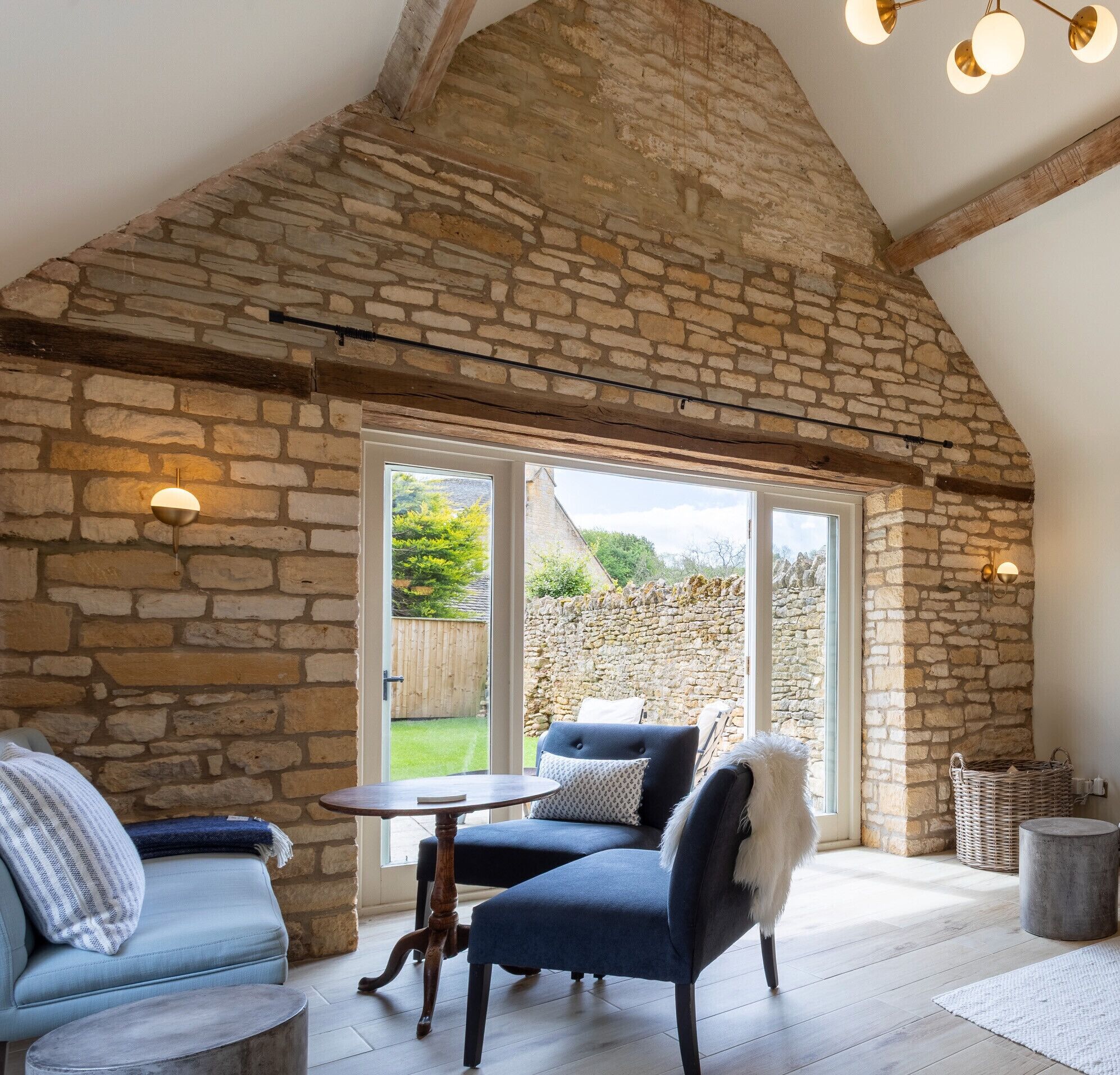 A cozy room with stone walls, a wooden ceiling, and a large window. There is a blue sofa, a small round table, and a wicker basket.