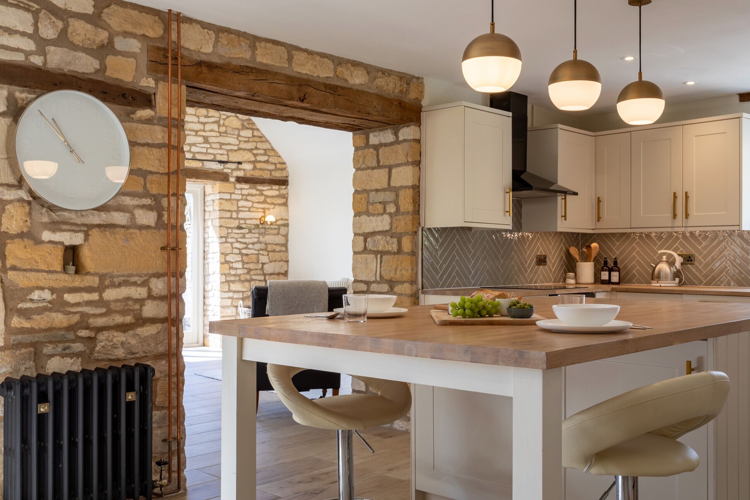 Una cocina con pared de piedra, vigas de madera, un reloj redondo, lámparas colgantes y una barra con taburetes.