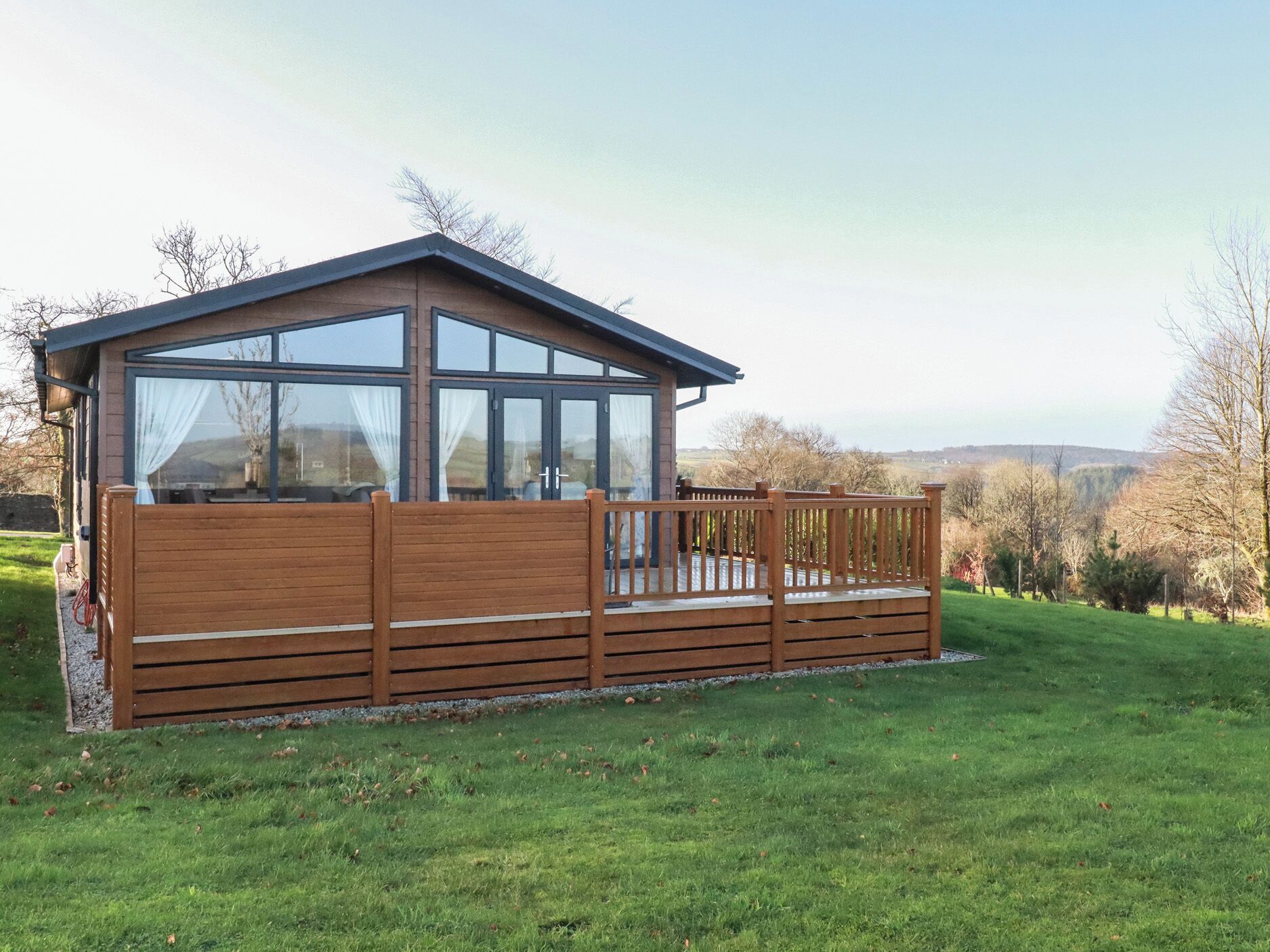 A wooden cabin with large windows and a deck, surrounded by a grassy area and trees.