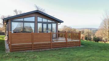 A wooden cabin with large windows and a deck, surrounded by a grassy area and trees.